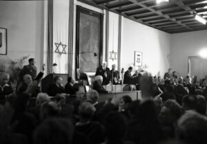 חברי האסיפה המכוננת מצביעים בישיבתה הראשונה, 1949. מאחורי הבמה דגלי ישראל ודיוקן הרצל